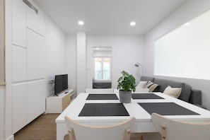 A modern, minimalist room with a white table, two chairs, a grey sofa, a potted plant, and a television on a side table.