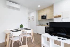 Une cuisine moderne avec des armoires blanches, un four à micro-ondes et un four encastré. On y trouve un coin repas avec une table et des chaises, ainsi qu’un téléviseur à écran plat posé sur un meuble blanc.