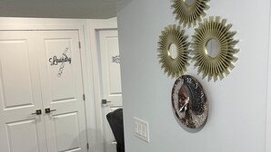 A hallway with a white door labeled "Laundry" and "Bath," a wall clock, and three sunburst mirrors. - Discover your ideal getaway. Relax and recharge in a space that feels like home. (Edmonton)