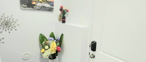 A hallway with a white door, a small shelf with decorative items, a wall-mounted picture, and a floral arrangement on a pedestal.