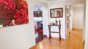 Un pasillo con suelo de baldosas, una zona de comedor con alfombra roja, una consola decorada con objetos y un cuadro grande con motivos florales en la pared.