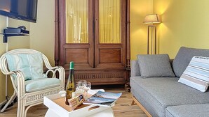A living room with a wooden wardrobe, a grey sofa, a wicker chair, a white coffee table with a bottle, glasses, and a magazine.