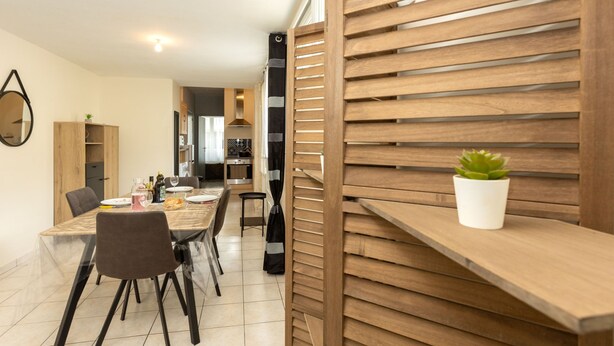 A modern dining area with a wooden table and chairs, a kitchen in the background, and a potted plant on a shelf.