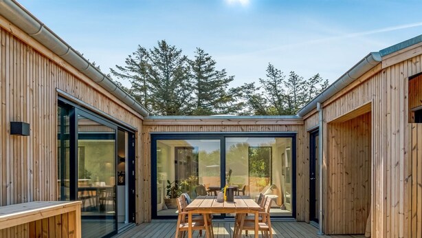 A wooden deck with a table and chairs, surrounded by glass doors and windows, overlooking a forested area.