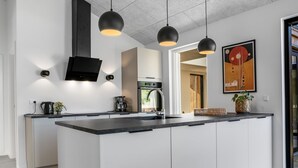 A modern kitchen with a large island, black pendant lights, a flat-screen TV, and a framed artwork on the wall.