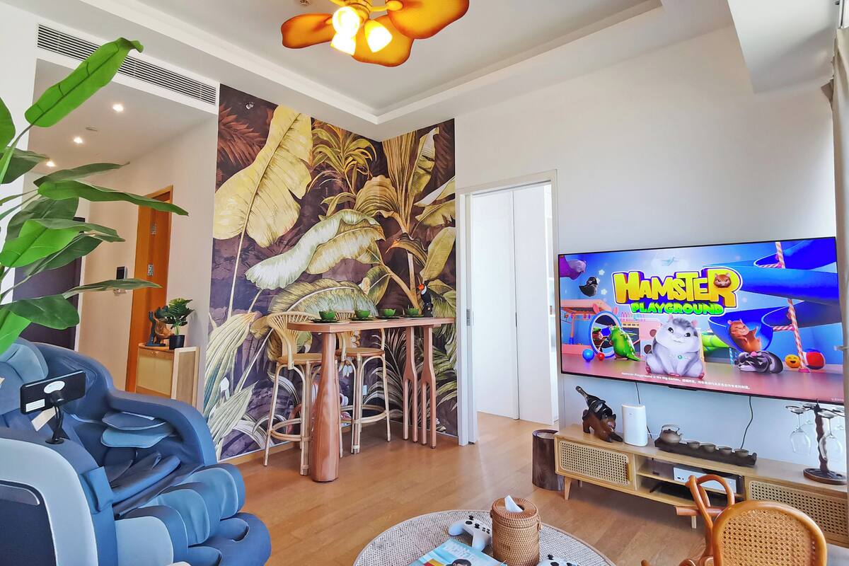A living room with a television displaying 'Hamster Playground', a ceiling fan, a dining table with chairs, and a large plant.