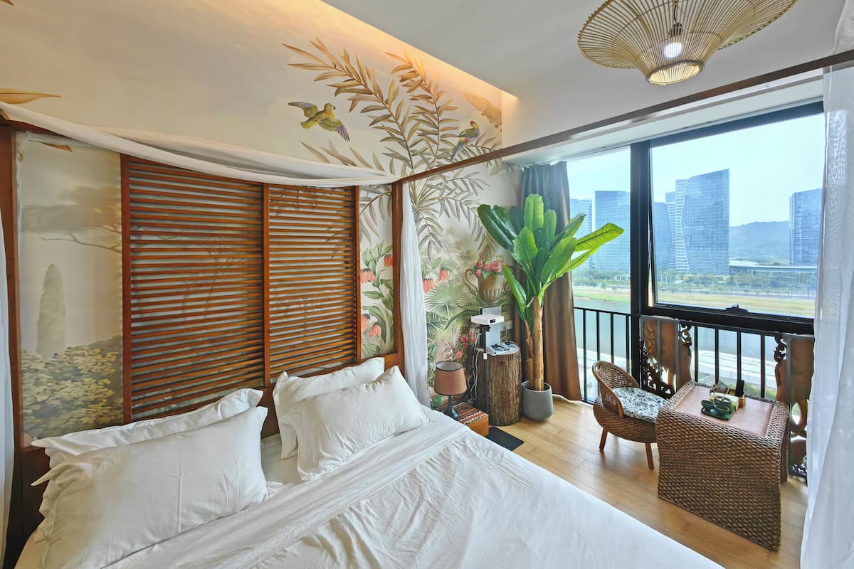 A modern hotel room with a large bed, a balcony with wicker furniture, and a view of city buildings.