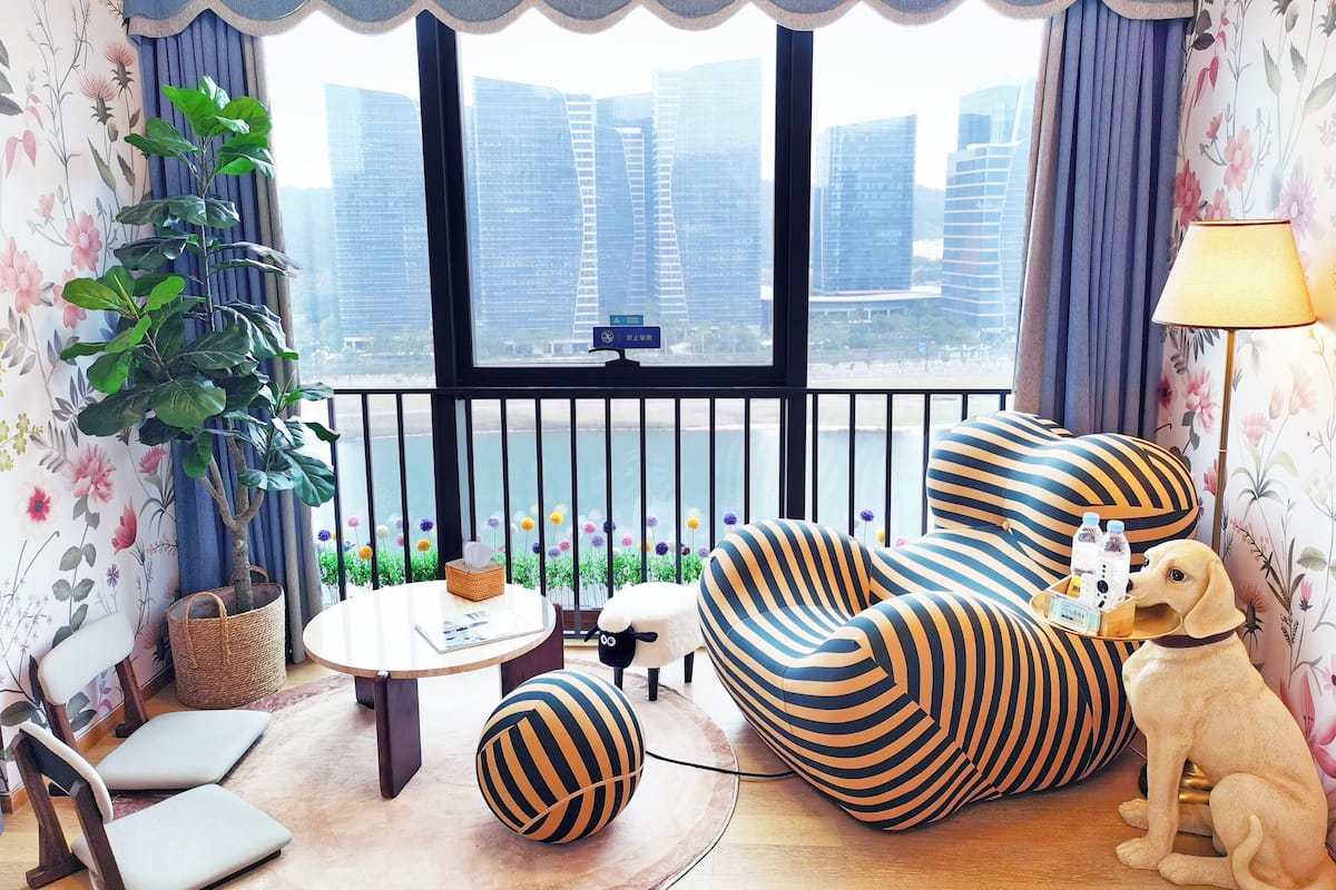 A modern living room with striped sofas, a dog, and a view of tall buildings through large windows.