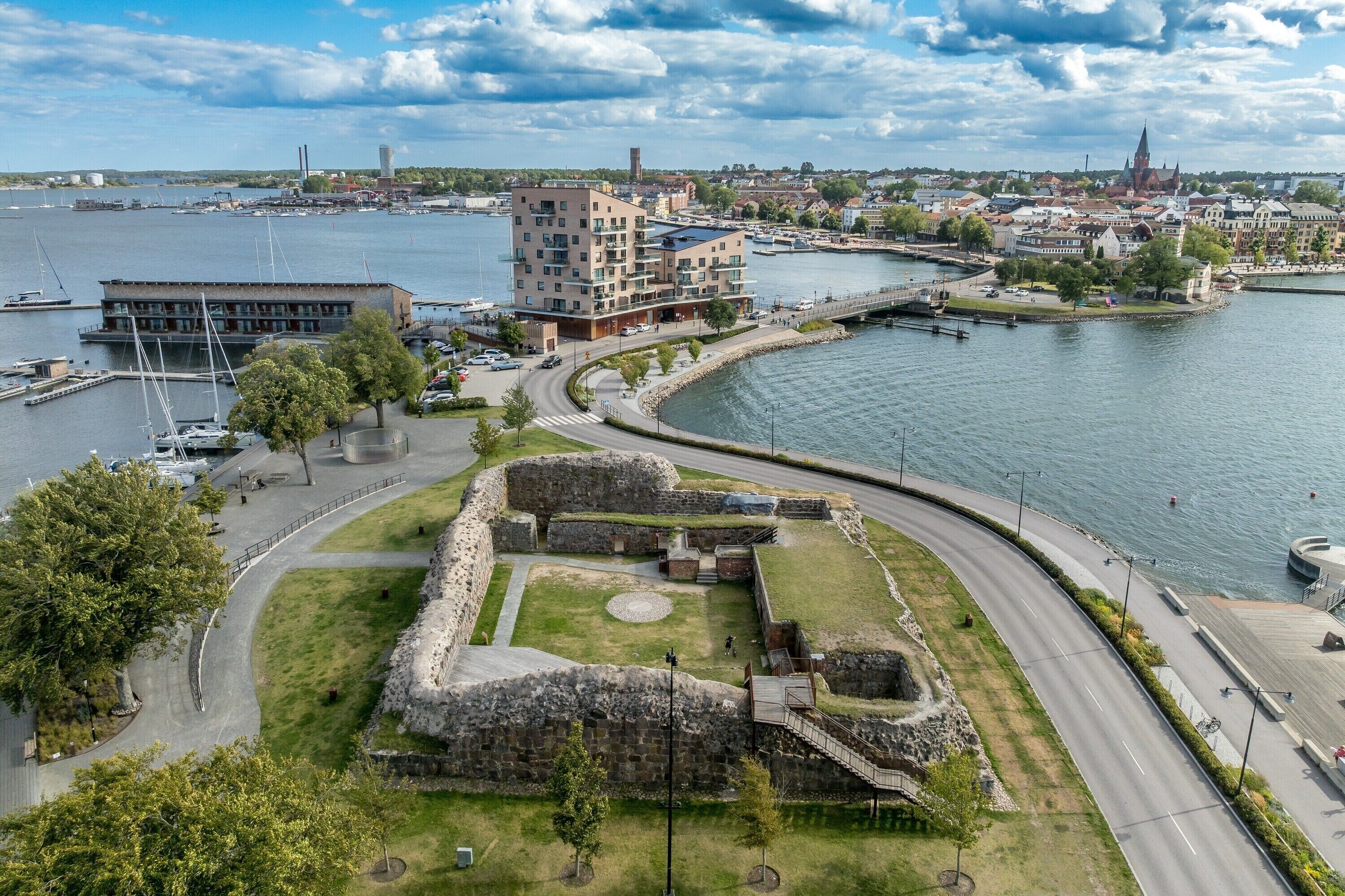 En fastighet vid vattnet med en stenfästning, moderna byggnader och en småbåtshamn.