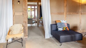 Ein Zimmer mit Holzboden, einer schwarzen Couch mit gemusterten Kissen, einem flauschigen Sessel und einem weißen Vorhang.