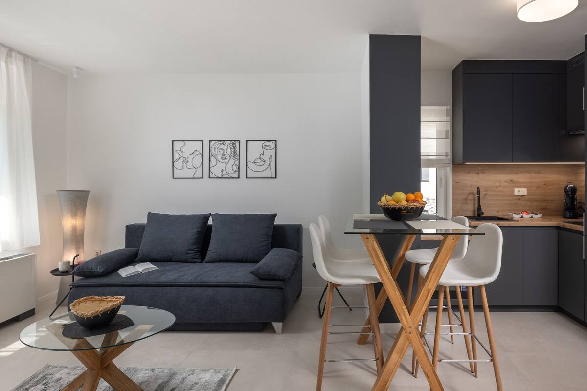 Una sala moderna con un sofá oscuro, una mesa de centro de vidrio y una zona de comedor con sillas blancas y una mesa de madera.
