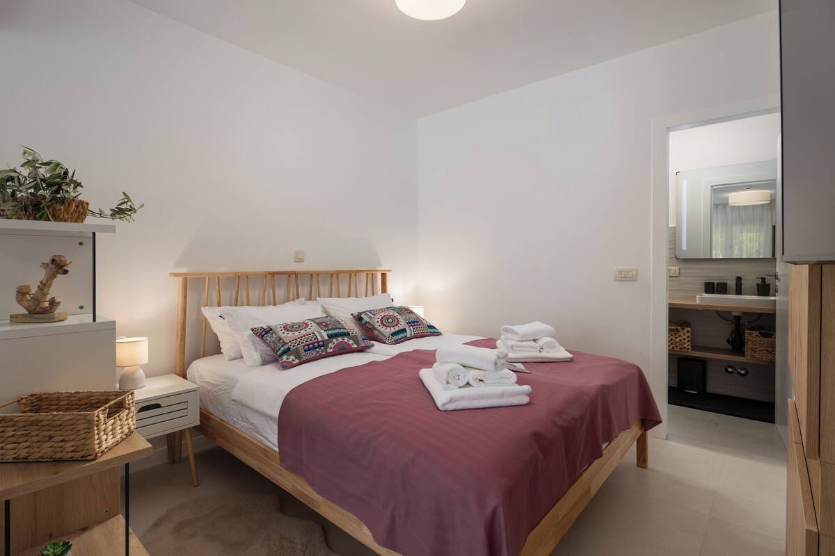 Un dormitorio ordenado con cama, mesita de noche, lámpara y una planta pequeña.