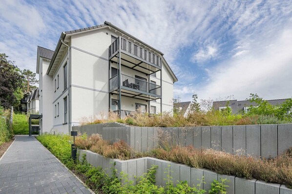 A modern multi-story residential building with a balcony, a paved walkway, and a landscaped area with plants.