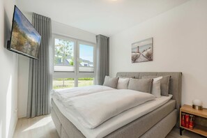 A bedroom with a bed, a TV mounted on the wall, a window with curtains, and a painting on the wall.