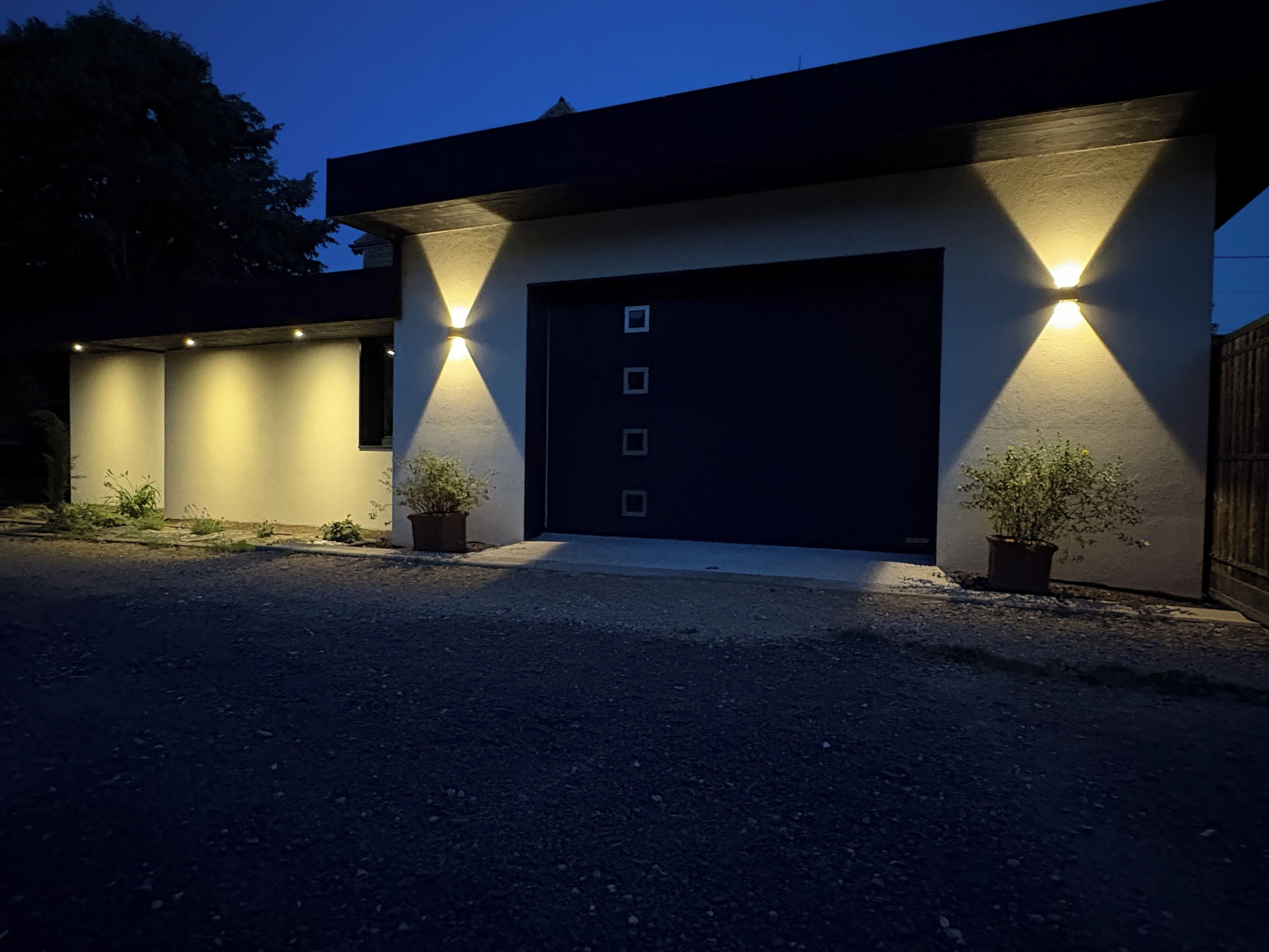 Une maison moderne avec une porte de garage sombre, deux plantes en pot et un éclairage extérieur.