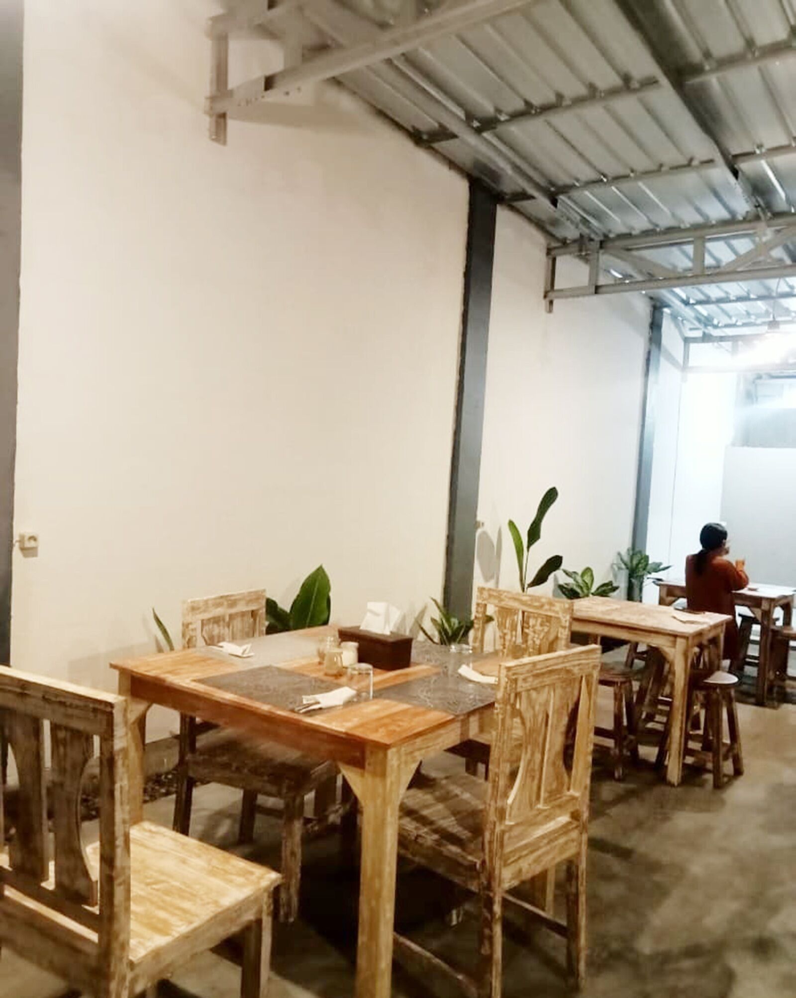A dining area with wooden tables and chairs, potted plants, and a person seated at a table.