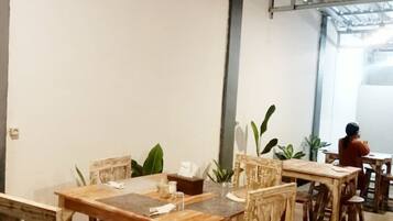 A dining area with wooden tables and chairs, potted plants, and a person seated at a table.