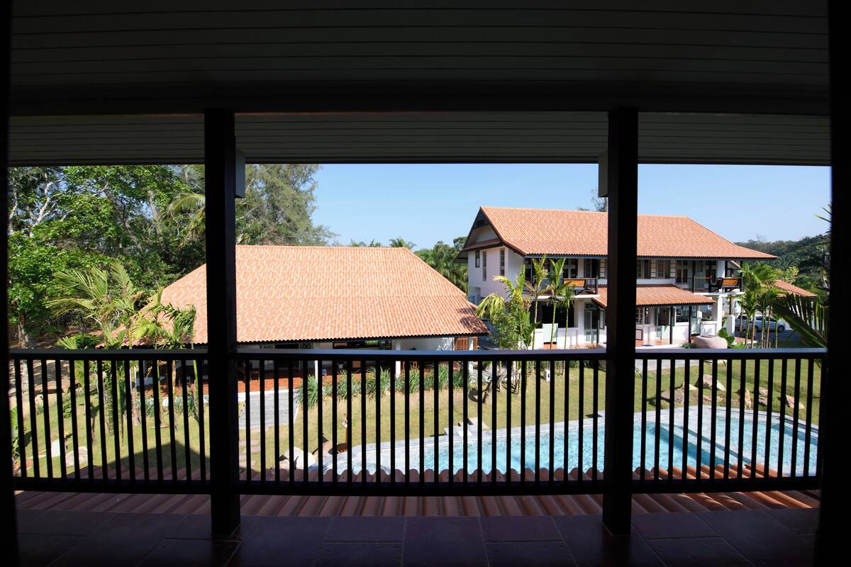 A view from a covered balcony with a pool, featuring a house with a tiled roof and a well-maintained lawn.