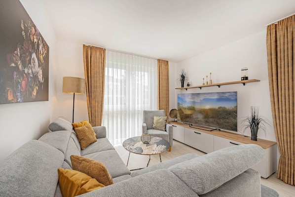 A modern living room with a grey sofa, a round coffee table, a TV on a wooden stand, and a large window with sheer curtains.