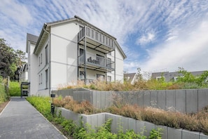 A modern multi-story residential building with balconies, a paved walkway, and surrounding greenery.