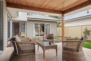 A covered patio area with wicker furniture, a glass table, and a view of a house with sliding glass doors. - Leeuwin House—Spacious Seaside Living for Groups (Quindalup)