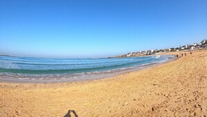 A wide sandy beach with gentle waves, a clear blue sky, and a few buildings in the background.