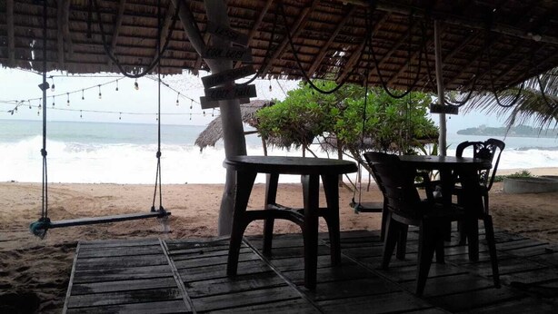 A beachside seating area with a swing, a table, and chairs under a thatched roof, overlooking the ocean. - Mami Beach Resort (Tangalle)