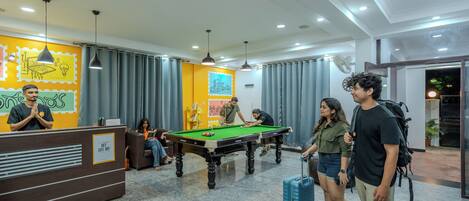 A pool table in a hotel lobby with a reception desk and a fan on the wall.