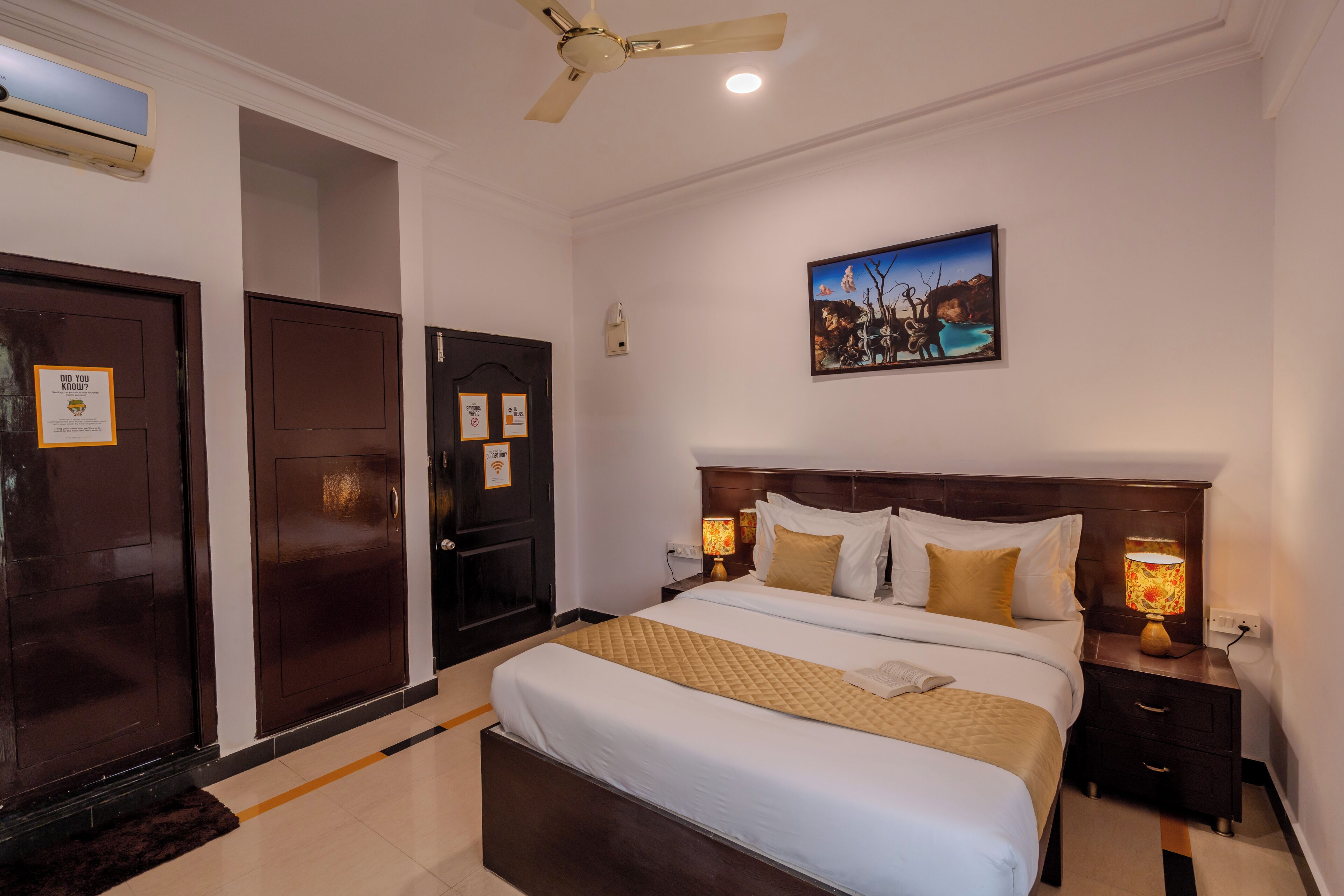 A hotel room with a large bed, a nightstand with a lamp, a ceiling fan, and a painting on the wall.