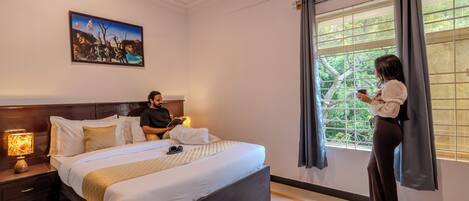 A hotel room with a large bed, a nightstand with a lamp, a painting on the wall, and a window with curtains.