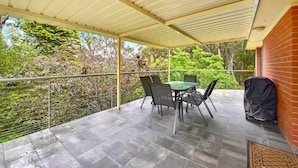 A covered patio with a table and chairs, a grill, and a view of greenery.