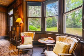 Quarto aconchegante com paredes de madeira, uma janela grande, duas cadeiras, uma mesinha com um tabuleiro de xadrez e uma estante de livros.