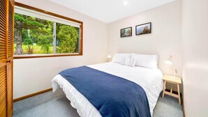 Quarto com uma grande janela, uma cama com roupa de cama branca, uma mesa de cabeceira com abajur e duas molduras na parede.