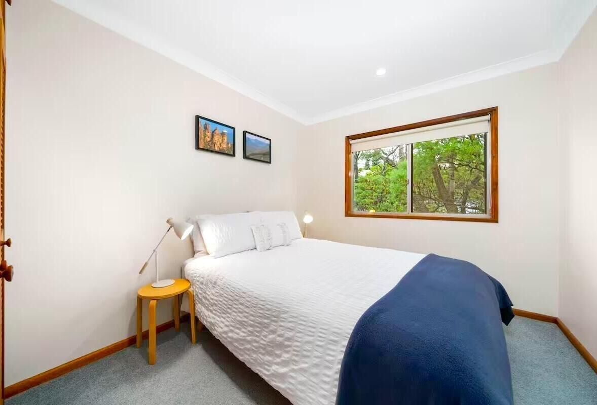 A bedroom with a bed, a nightstand, a lamp, a window with a view of trees, and two framed pictures on the wall.