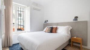 A neatly made bed with white bedding, a small wooden nightstand, and a window with curtains.