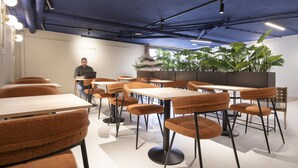 A modern lounge area with multiple tables and chairs, a person seated at a table, and a large potted plant in the background.