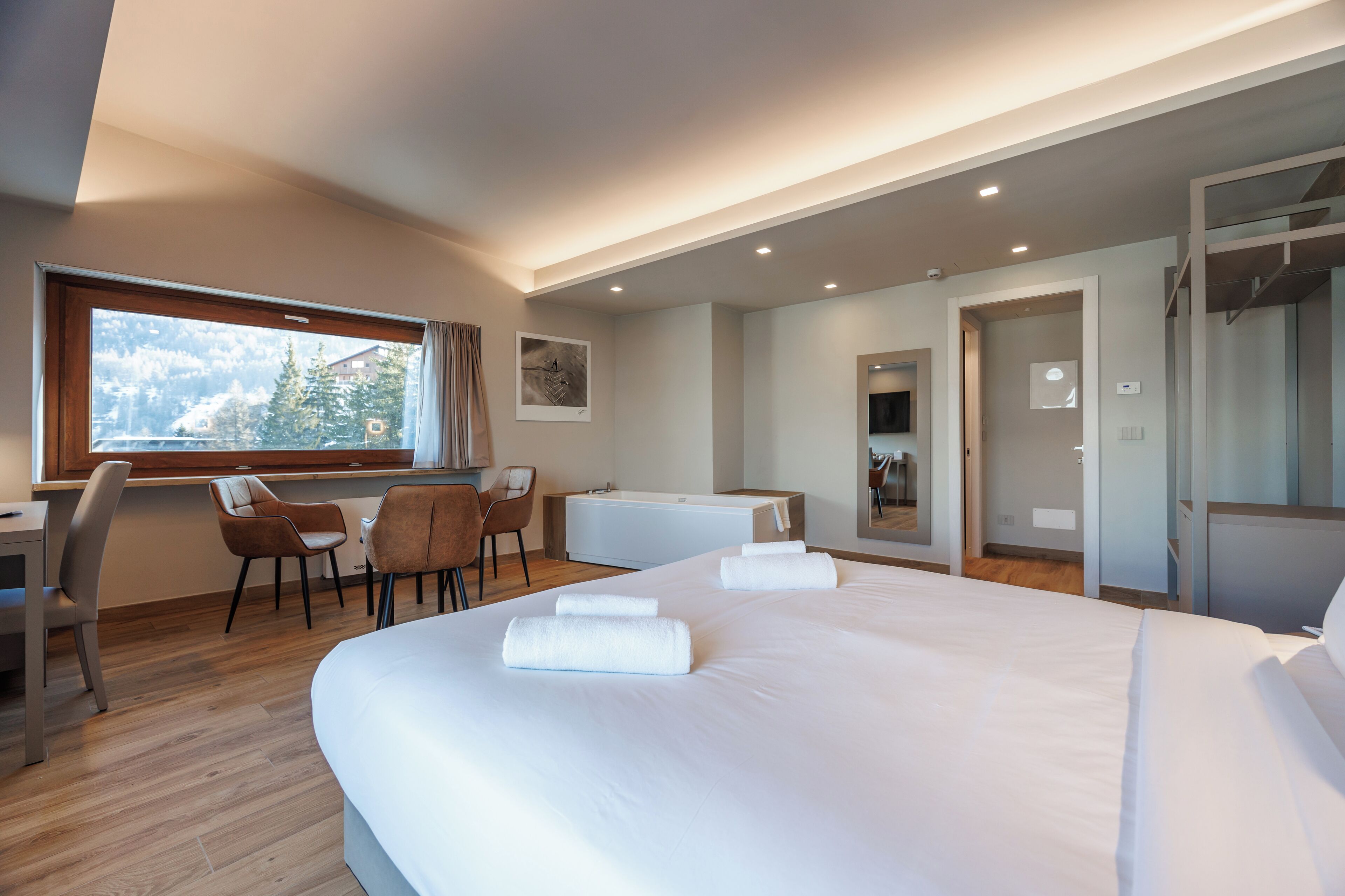 Habitación de hotel moderna con una cama grande, un escritorio y una silla. Hay una ventana que ofrece vistas a árboles y una montaña.