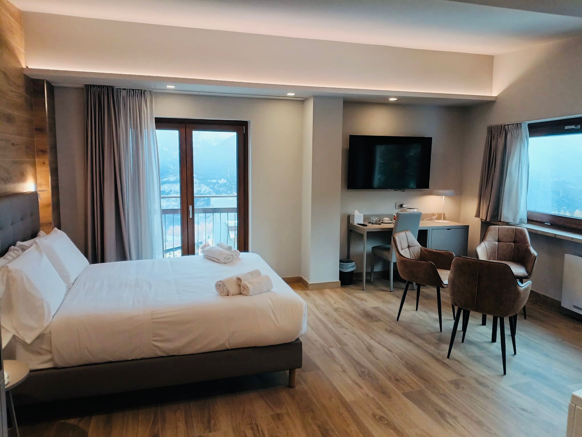Une chambre d’hôtel avec un grand lit, un bureau avec une chaise, une télévision et un balcon avec des rideaux.