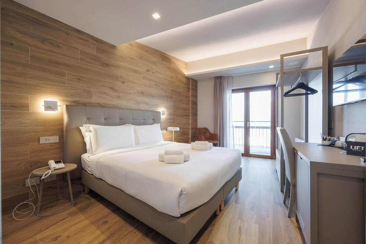 Ein modernes Hotelzimmer mit einer Holzwand, einem großen Bett mit weißen Leinen, einem Nachttisch mit Telefon sowie einem Schreibtisch mit Spiegel und Kleiderhaken.