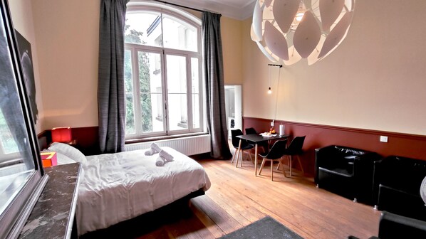 A hotel room with a bed, a desk, a chair, a black sofa, a window with curtains, and a ceiling light fixture.