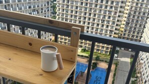 A balcony with a wooden bench and a white mug. In the background, there are high-rise buildings and a swimming pool with the word "SAEKYUNG" visible. - Nicosia's Place (Lapu-Lapu)