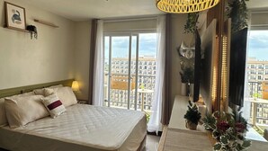 A modern hotel room with a large bed, a desk with a laptop, a view of the city through the window, and a decorative plant. - Nicosia's Place (Lapu-Lapu)