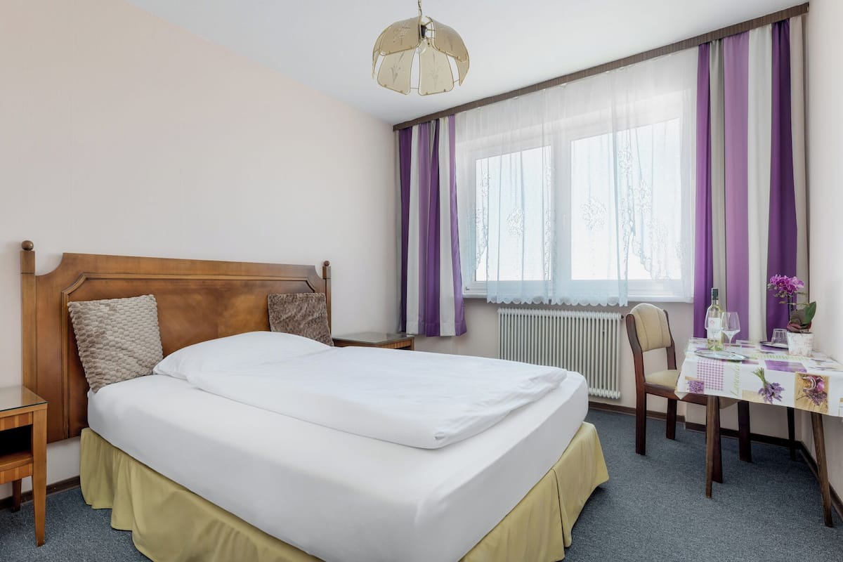 A hotel room with a large bed, a wooden headboard, a bedside table, a chair, a small table with a vase of flowers, and a window with purple curtains.