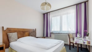 A hotel room with a large bed, a wooden headboard, a bedside table, a chair, a small table with a vase of flowers, and a window with purple curtains.