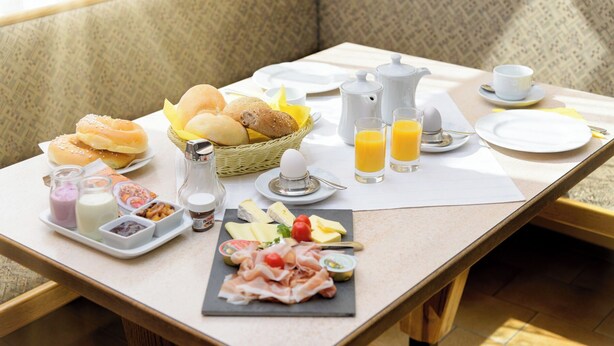 A breakfast setup with bread, pastries, cheese, meats, and juice on a table in a room with patterned walls.