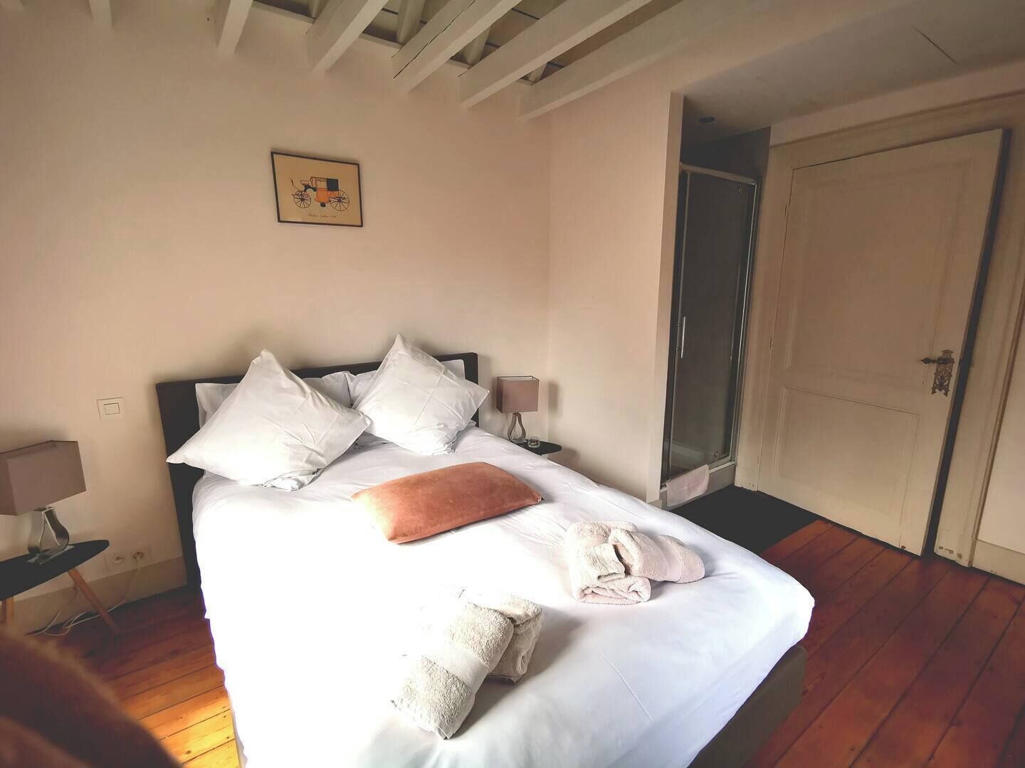 Ein ordentlich bezogenes Bett mit weißen Leinen und Kissen, ein Nachttisch mit Lampe, ein gerahmtes Bild an der Wand und eine Tür zu einem Duschbereich.