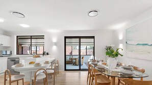 A modern kitchen with white cabinets, a central island, and a dining area with a glass table and wooden chairs. - Solace Jervis Bay - 3BR relaxed beach side living (Vincentia)