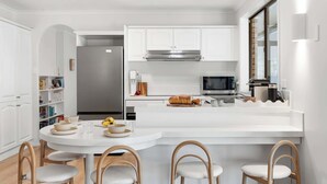 A modern kitchen with white cabinetry, a central island with seating, a microwave, and a refrigerator. - Solace Jervis Bay - 3BR relaxed beach side living (Vincentia)