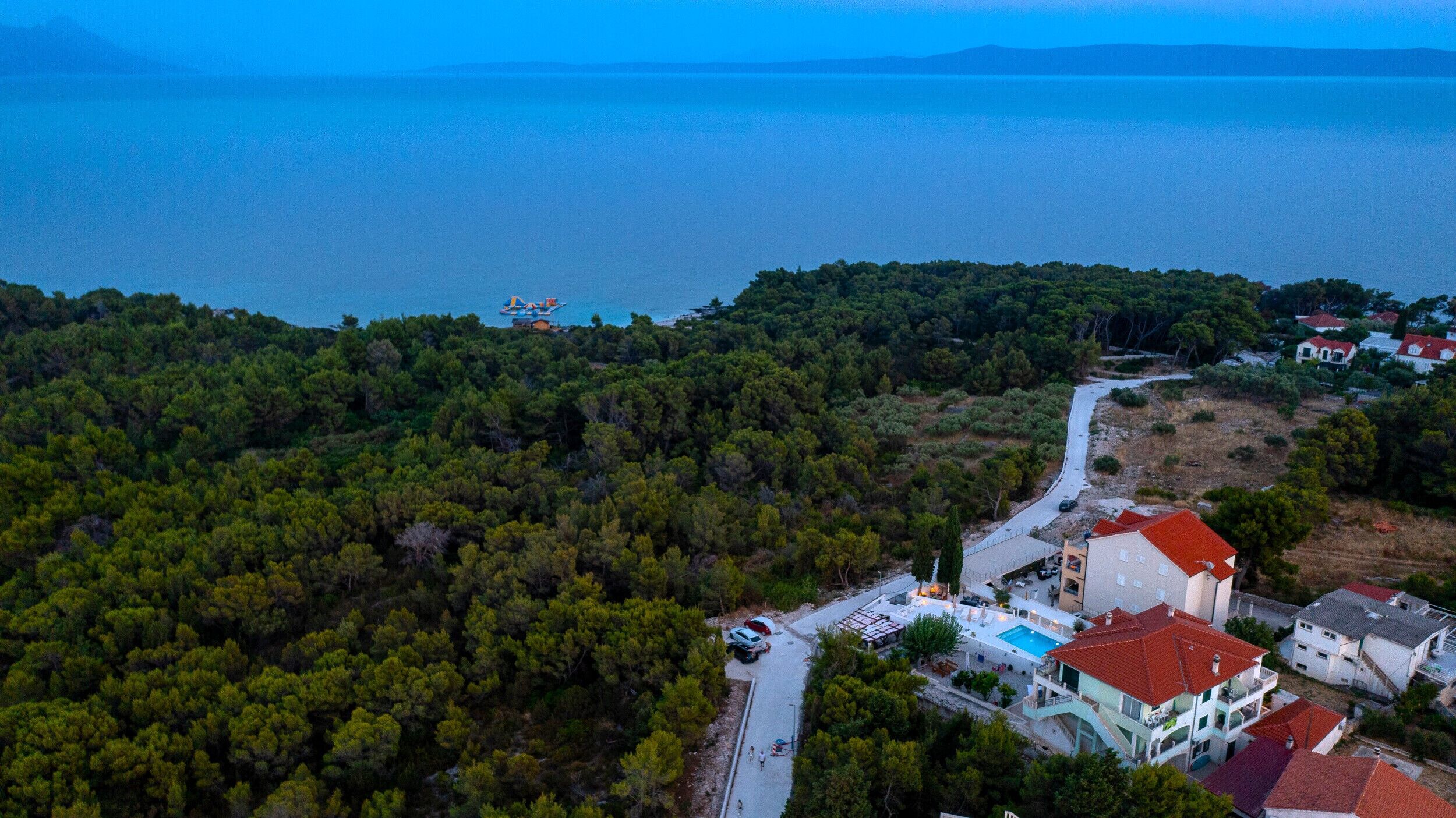 An aerial view of a coastal area with dense forest, a road, and buildings with red roofs.