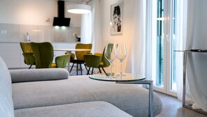 A modern living room with a grey sofa, a glass coffee table, and two wine glasses. In the background, there is a dining area with green chairs and a white table.
